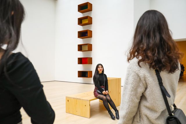 a woman sitting on a bench in front of a donald judd piece on the wall, made of squares suspended on the wall, each a different color